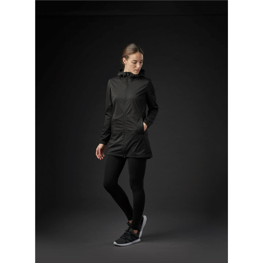 A woman wearing a black softshell jacket, black leggings, and athletic shoes, standing against a dark background.