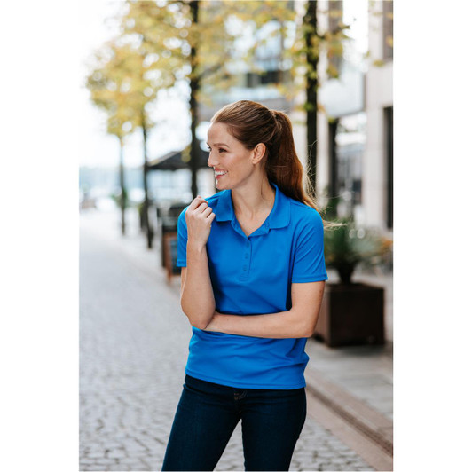 A woman wearing a blue polo shirt stands outdoors, smiling and looking to the side, with a modern urban setting in the background.