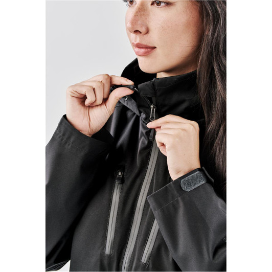 A woman in a black lightweight rain jacket adjusts the zip and hood, featuring a logo on the sleeve.
