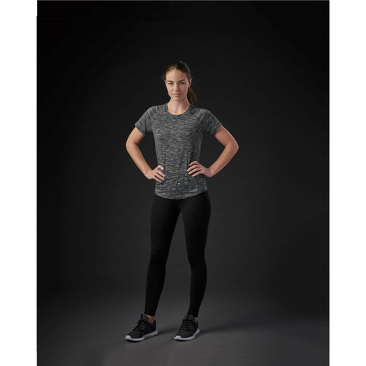 A woman poses in a fitted, multicoloured grey Pacifica tee, paired with black leggings and shoes against a dark background.