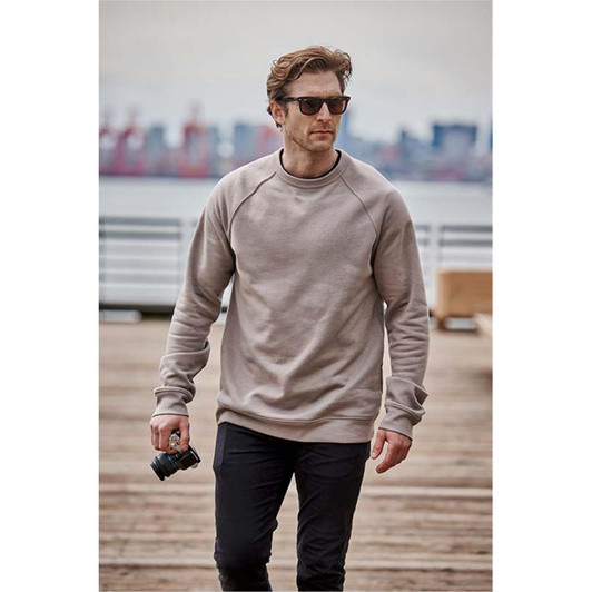 A man wearing a beige crew neck fleece sweater and sunglasses, holding a camera while walking on a pier.