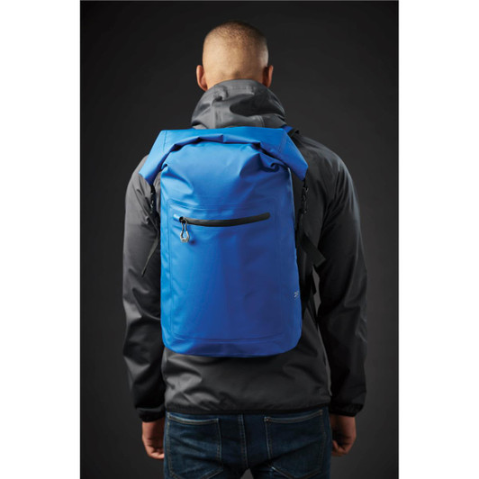 A blue backpack is worn on a person's back, featuring a front zipper pocket. The person is dressed in a black jacket.