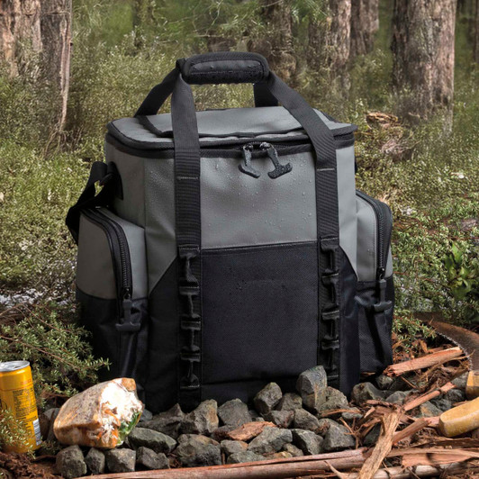 A grey and black cooler bag sits among rocks and foliage, featuring multiple compartments and a sturdy handle.