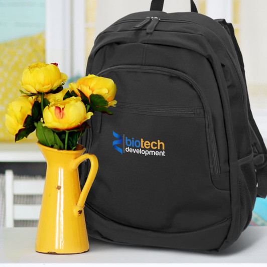A black backpack with a logo and a front pocket, placed next to a yellow vase with flowers.
