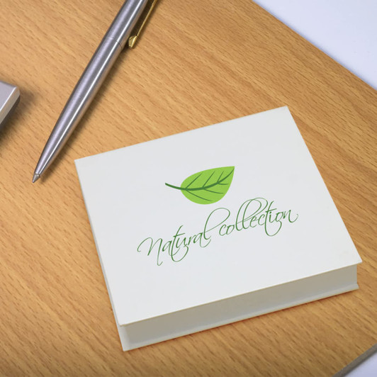 A square sticky note pad in cream with a green leaf design, resting on a wooden surface beside a pen.