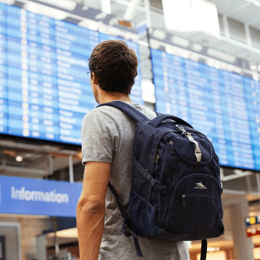 A navy blue backpack worn by a person facing an airport information board. The backpack has a logo and multiple compartments.
