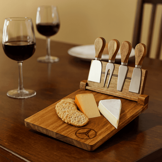 A wooden cheeseboard set featuring four stainless steel knives, cheese, and crackers, with two glasses of red wine in the background.