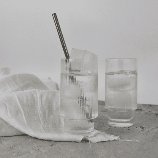 Two ribbed highball glasses filled with clear liquid and ice, alongside a white linen cloth and a metal straw.