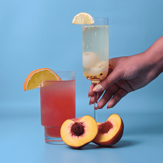 A set of ribbed highball glasses containing brightly coloured drinks, with peaches and lemon slices in the foreground.
