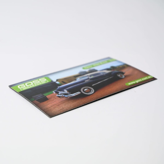 A square cut fridge magnet featuring a vintage black car on a dirt road with a logo in the corner.