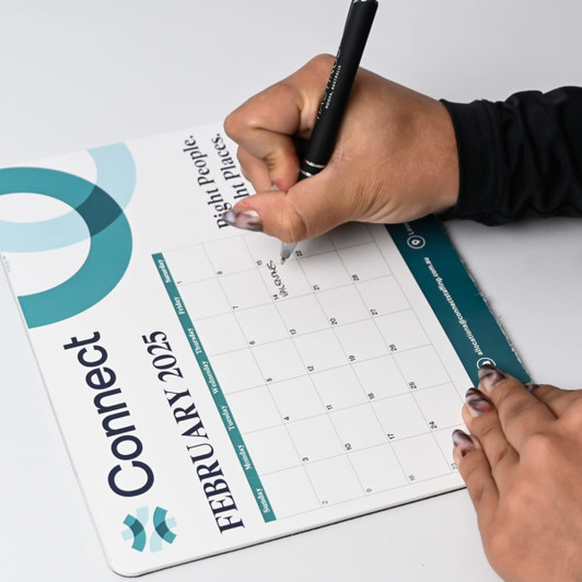 A mouse mat featuring a calendar for February 2025, with a teal and white design, and a person writing on it.