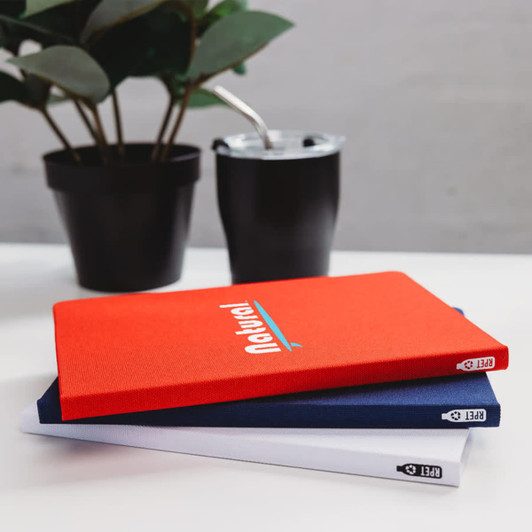 Three A5 notebooks in red, white, and navy stacked on a table with a plant and a drink bottle in the background.
