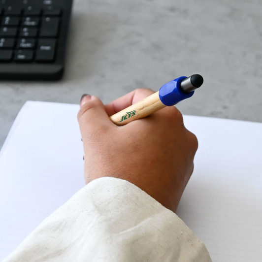 A bamboo spinner pen with a blue grip in a hand, resting on a lined notebook with a keyboard nearby.