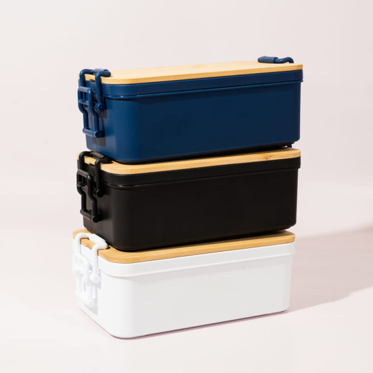 Three stacked lunch boxes in navy blue, black, and white, each with a wooden lid and a branded logo.