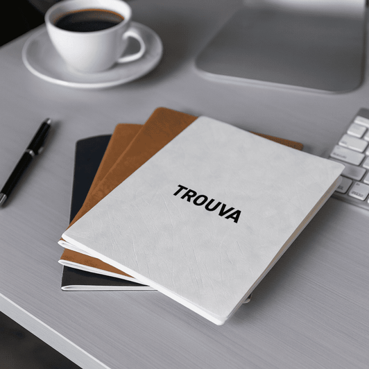 A set of three A5 notebooks in white, black, and brown, featuring a logo, placed on a desk with a coffee cup.