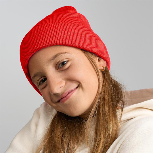 A red knitted beanie worn by a child with long hair, featuring a logo on the side.