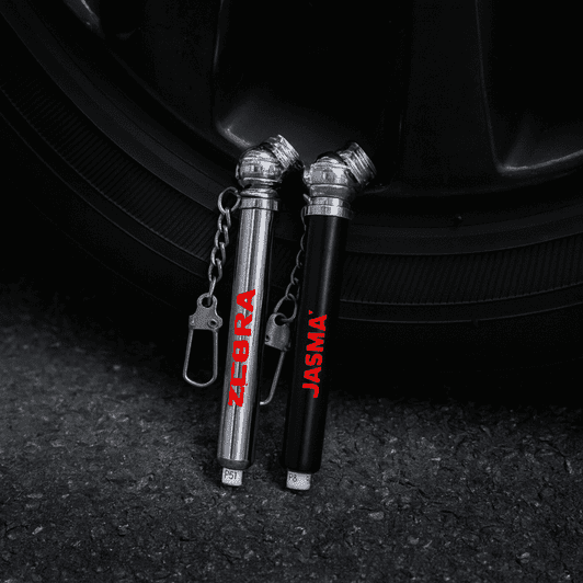 Two key chain tyre gauges, one silver and one black, attached to a keyring, positioned near a tyre.