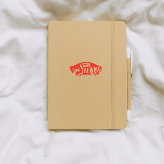 A tan A5 notebook with a logo, featuring a pen attached, resting on a light fabric background.