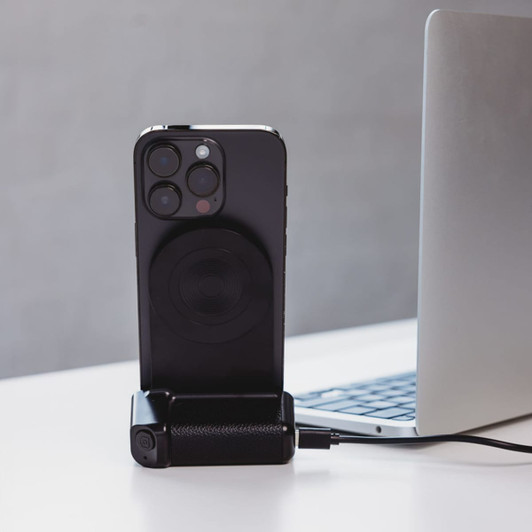 A black wireless charger with a phone attached, placed next to a laptop on a white surface.