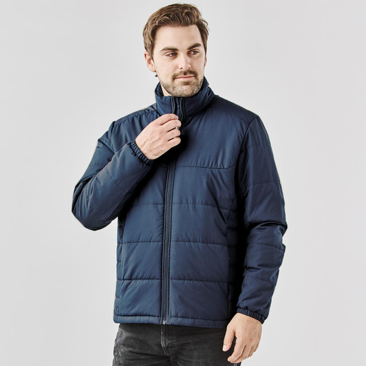 A male model wearing a navy blue puffer jacket with a high collar and zip closure. The jacket has an elasticised hem.
