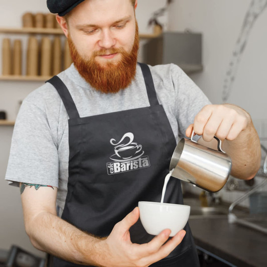 A man with a beard in a black Savoy bib apron pours milk from a metal jug into a white bowl.