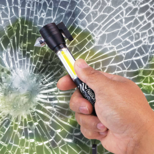 A black vehicle safety tool with a light, held in a hand, against a backdrop of shattered glass.