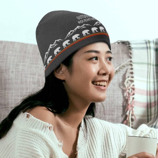 A grey custom knitted beanie with a mountain and bear design, worn by a smiling person holding a drink.