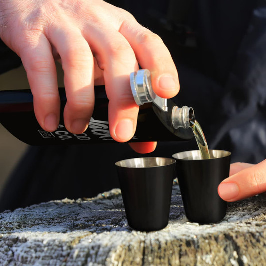 A drink bottle being poured into two black stainless steel shot glasses, set on a wooden surface.