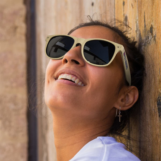 Malibu Basic Sunglasses in natural colour, featuring a logo, worn by a smiling person against a rustic wooden background.