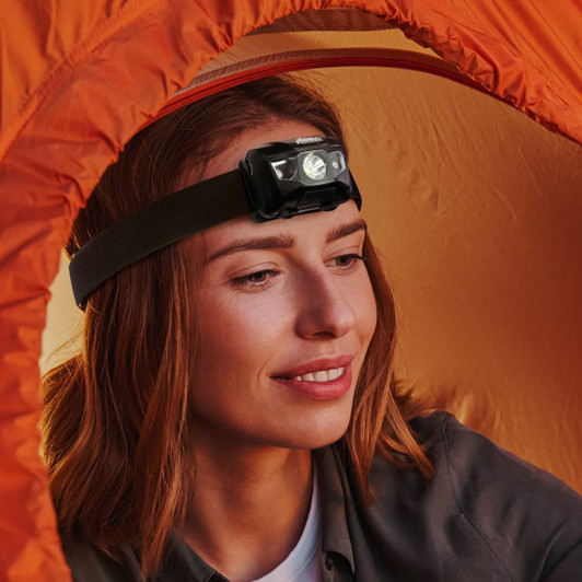 A black headlamp torch worn by a woman, set against an orange tent background. The product has a sleek design.