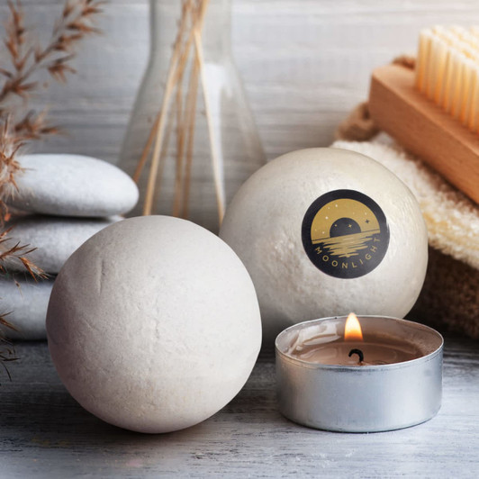 Two round, pale bath bombs sit beside a small candle, with smooth stones and a wooden brush in the background.