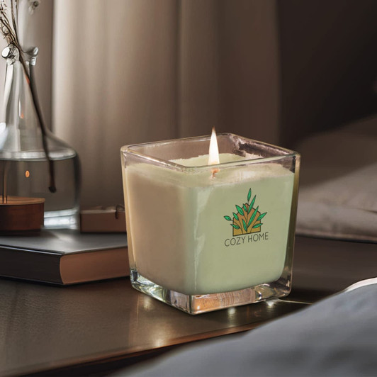 A green, square scented candle in a glass container with a logo, placed on a bedside table beside a book.