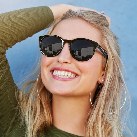 A pair of stylish black sunglasses with a classic frame, worn by a smiling woman with long blonde hair.