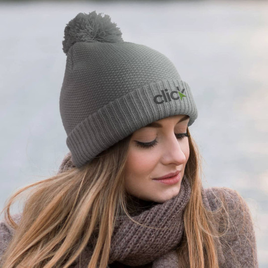 A grey knitted beanie featuring a pom-pom on top and a logo, worn by a woman with long hair.