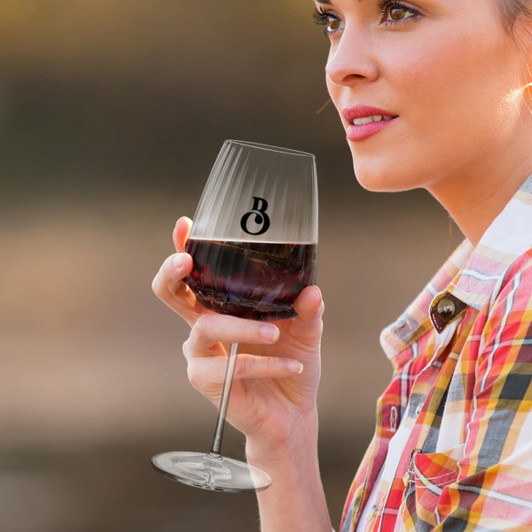 A woman holds a clear wine glass filled with red wine, featuring a logo on the side. The glass has a stem and a curved bowl.