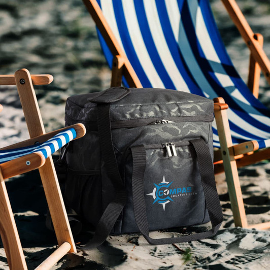 A grey camo cooler bag sits next to striped beach chairs on sand, featuring a logo.
