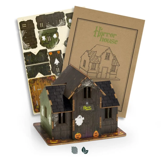 A wooden model of a haunted house in dark colours, featuring a ghost and pumpkins, with assembly instructions.