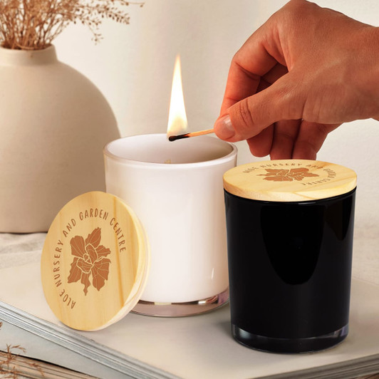 A white and black scented candle with wooden lids, one being lit with a match, beside some dried flowers.