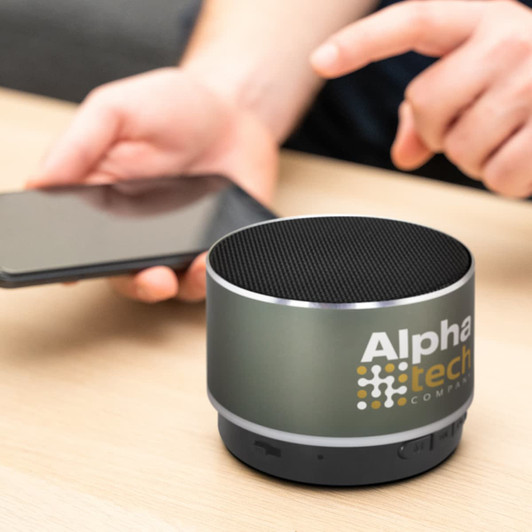 A Bluetooth speaker in grey with a mesh top, featuring a logo, positioned on a table next to a smartphone.