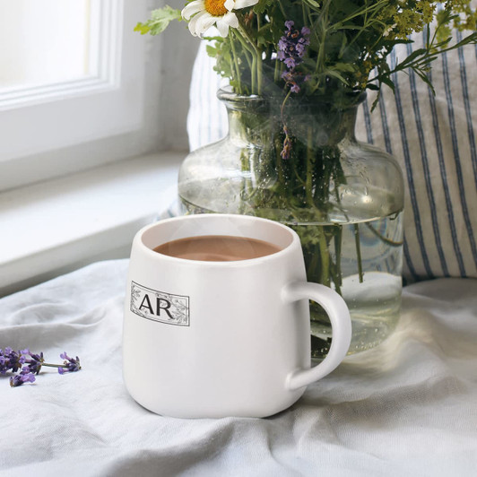 A white coffee mug with a handle, filled with coffee, beside a vase of flowers on a linen surface.