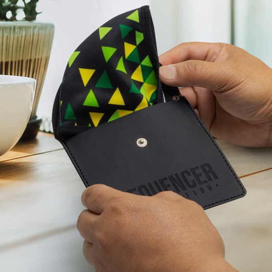 A black key ring pouch with a colourful triangular pattern inside, held in a person's hands near a plant and cup.