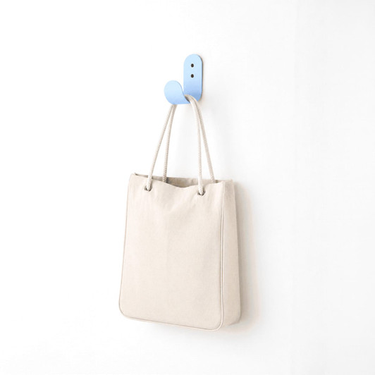 A cream-coloured tote bag hanging on a blue hook against a white wall. The bag has a simple design and a logo.