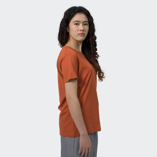 A woman in a rust-coloured T-shirt stands sideways, with curly hair and a neutral expression. The shirt features a logo.
