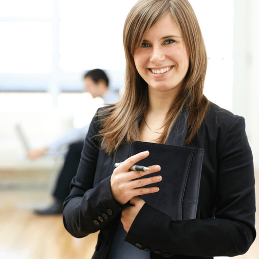 A young woman in a black blazer holds a dark padfolio and a pen, smiling in a bright office setting.