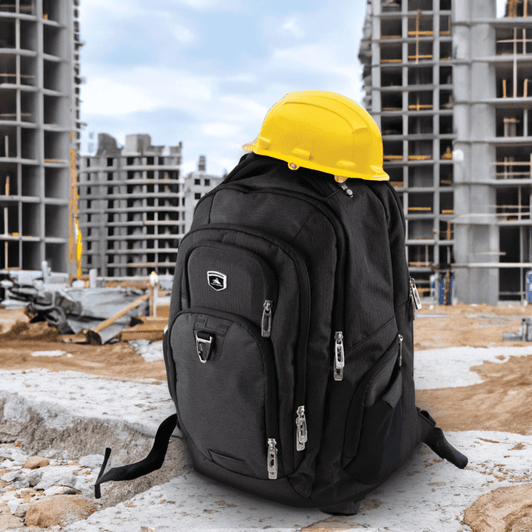 A black and grey 42L computer backpack with multiple compartments, placed on a construction site beside a yellow hard hat.
