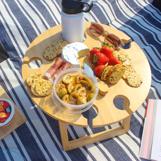 A wooden picnic table with snacks, fruit, a drink bottle, and sunglasses on a striped blanket.