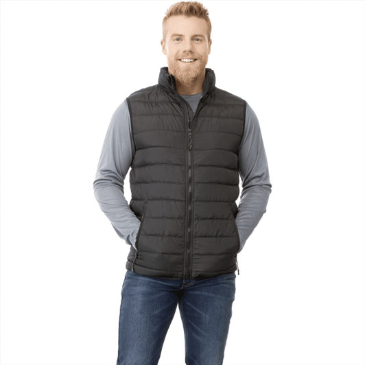 A man wearing a black insulated vest over a grey long-sleeve shirt, smiling and posing. The vest features a logo.
