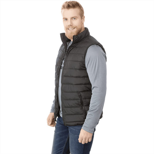 A man wearing a black insulated vest over a grey long-sleeve shirt, smiling and standing in profile. The vest has a logo.