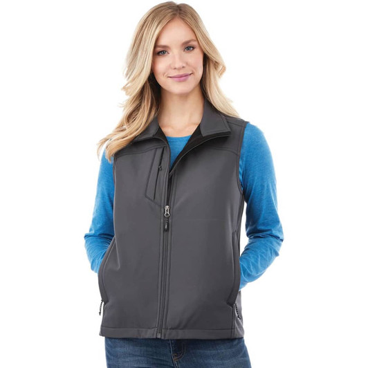 A women's softshell vest in dark grey, featuring a zip front and a logo. Model wears a blue long-sleeve top underneath.