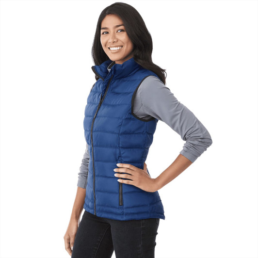 A woman wearing a blue insulated vest, styled with a grey long-sleeve top, smiling against a white background.